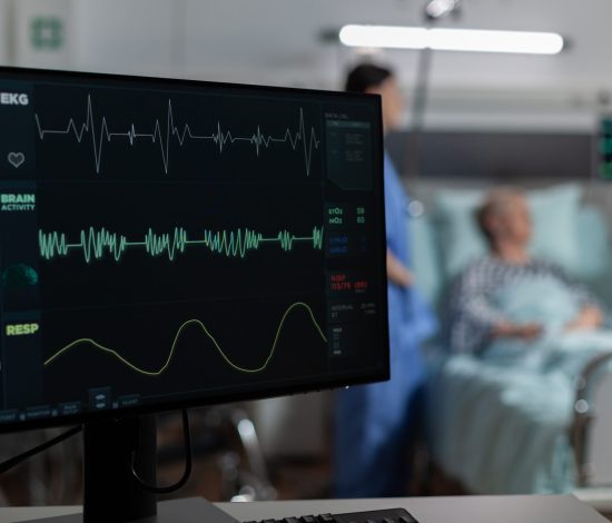Monitor in hospital ward showing bmp from patient, laying in bed with oxymeter attached on finger, discussing with doctor and nurse about recovery treatment.